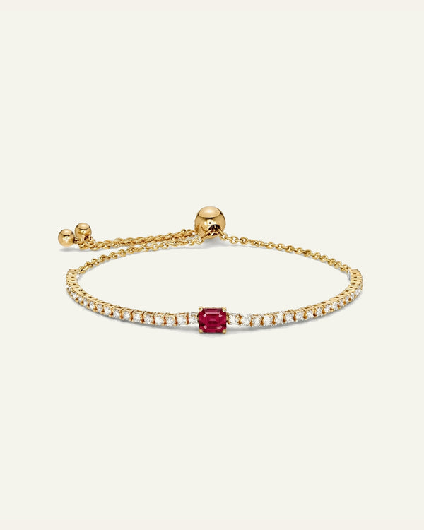 A delicate gold bracelet featuring a single emerald-cut red ruby center stone, flanked by rows of small clear pavé-set stones, displayed on a clean off-white background.