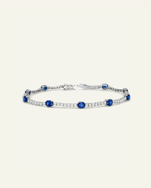 A silver tennis bracelet featuring round-cut blue sapphires and pavé-set clear stones, displayed on a solid cream background.