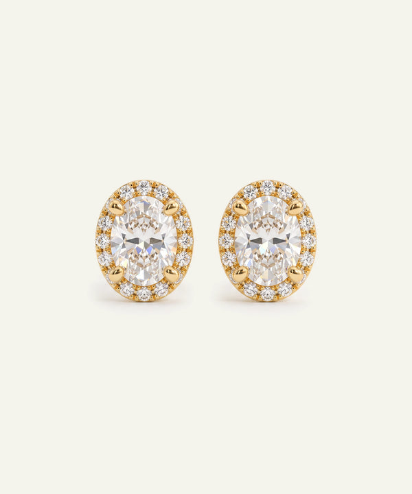A pair of oval stud earrings featuring large clear round-cut center stones surrounded by a halo of smaller clear stones in yellow gold settings, photographed on a clean off-white background.