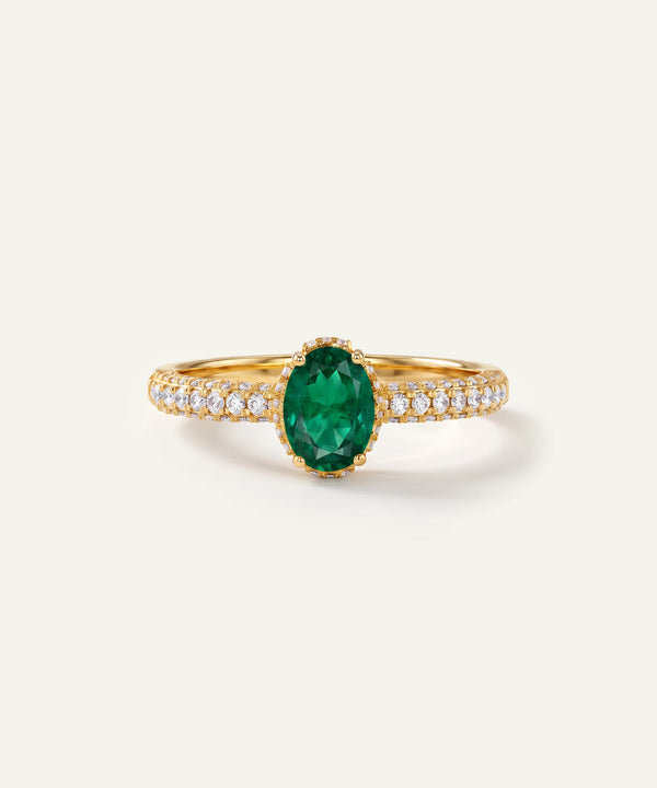 A yellow gold ring featuring a central oval-cut green emerald gemstone, flanked by pavé-set clear stones on the band, shown on a light cream background.