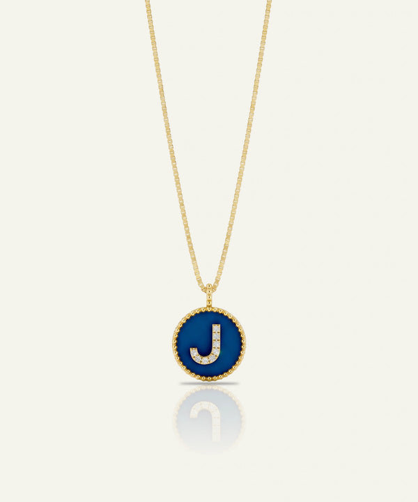 A gold chain necklace with a circular pendant featuring a deep blue enamel background and a raised gold letter 'J' outlined with small clear stones, shown on a light cream background with a soft reflection below.