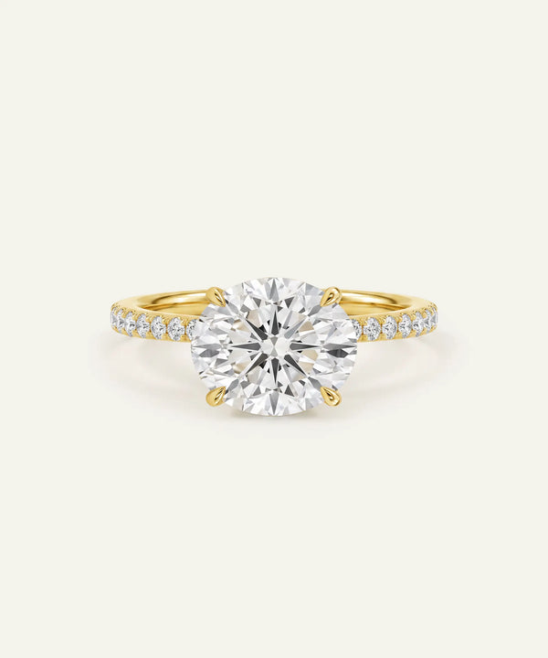 A yellow gold solitaire engagement ring featuring a large round brilliant-cut center stone and a pavé-set diamond band, shown on a light cream background.