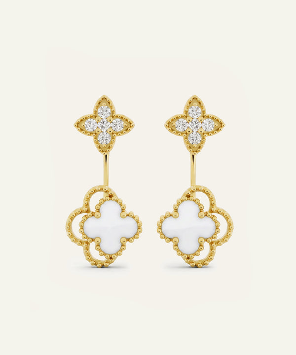 A pair of gold and white clover drop earrings with pavé-set clear stones on the top clovers and openwork gold clover shapes with white inlay on the bottom, shown on a light cream background.