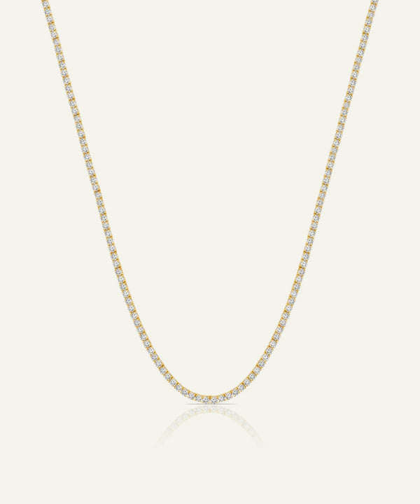 A delicate tennis-style necklace with small clear stones set in yellow gold, shown against a light cream background with a soft reflection below.