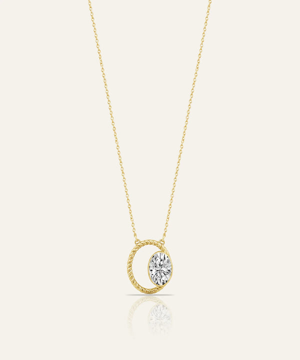 A delicate yellow gold chain necklace with a pendant featuring a twisted oval frame and a central bezel-set clear gemstone, shown on a light cream background with a soft reflection below.