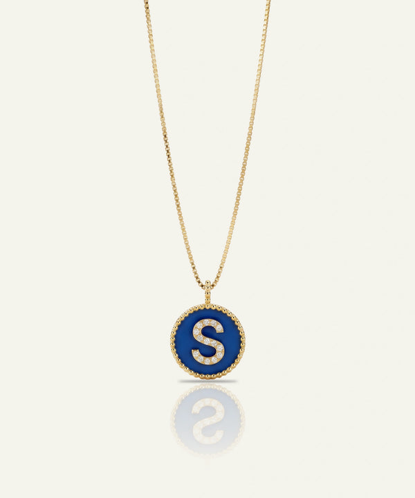 A gold chain necklace with a circular pendant featuring a deep blue enamel background and a white capital letter 'S' in the center, bordered by small clear stones, shown on a light cream background with a soft reflection below.