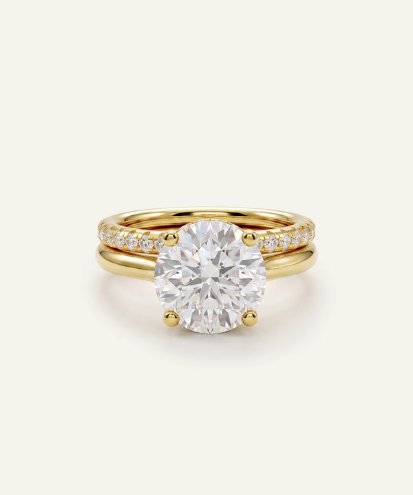 A yellow gold ring set featuring a large round-cut diamond solitaire ring with a pavé band and a slim matching gold wedding band behind it, set against a clean off-white background.