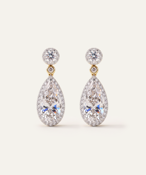 A pair of luxury teardrop dangle earrings featuring large pear-cut diamonds in a halo setting, connected to round brilliant-cut diamonds on gold-toned posts, set against a solid off-white background.