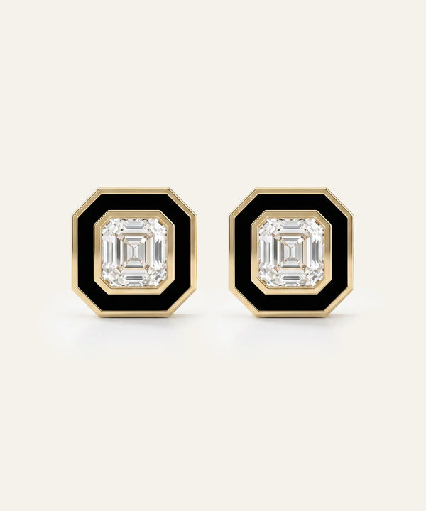 A pair of geometric stud earrings with emerald-cut clear stones, bordered by a black inlay and a gold octagonal frame, set against a light cream background.