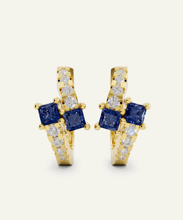 A pair of gold huggie hoop earrings featuring square blue sapphires and pavé-set clear stones, shown on a light cream background.
