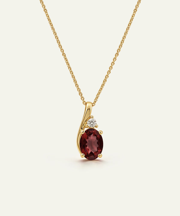 A gold pendant necklace featuring an oval deep red garnet stone and a small clear accent stone, displayed on a clean off-white background.