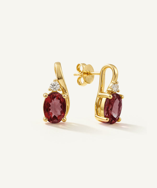A pair of gold post earrings featuring oval deep red gemstones and small clear accent stones, displayed on a clean off-white background.