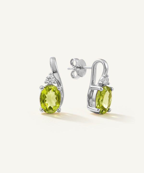 A pair of silver drop earrings featuring oval green peridot gemstones and small clear accent stones, shown on a clean off-white background.