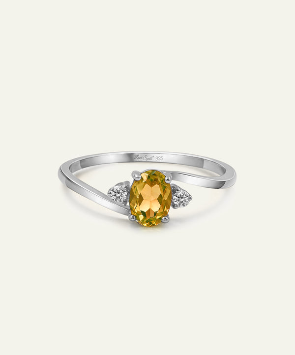 A silver ring featuring an oval-cut yellow citrine center stone, flanked by two small round-cut diamonds on a light cream background.