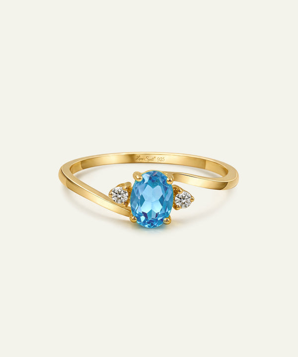 A gold ring featuring an oval-cut light blue gemstone in the center, flanked by two small round clear stones on a light cream background.