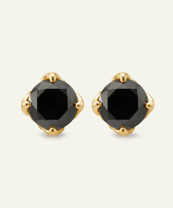 A pair of gold stud earrings featuring faceted black gemstones in a four-petal halo setting, shown against a light off-white background.