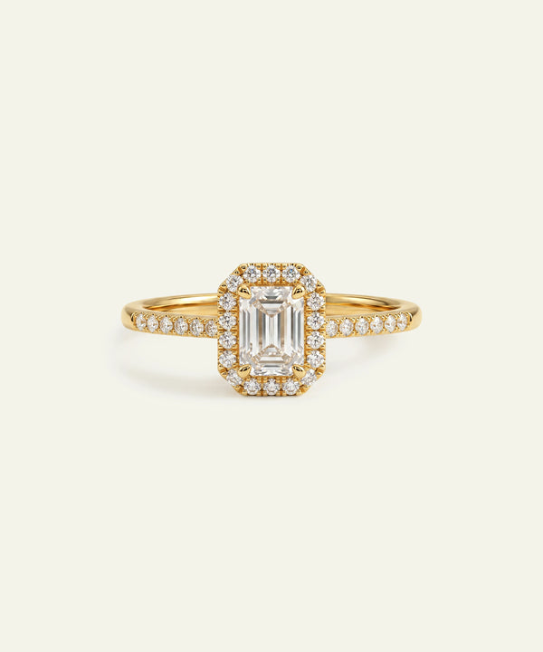 A yellow gold ring featuring a central emerald-cut clear gemstone surrounded by a halo of small round-cut stones, with a shank paved with similar stones, set against a solid off-white background.