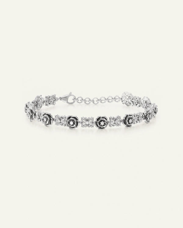 A silver chain bracelet featuring alternating rose-shaped links with dark center stones and clear diamond accents on a light cream background.