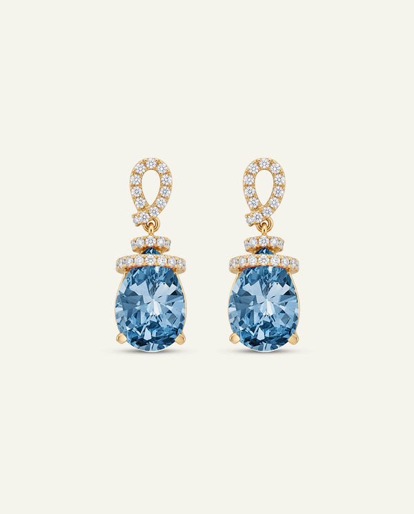 A pair of gold drop earrings featuring pear-cut light blue gemstones and pavé-set clear stones in the loops, shown on a light cream background.