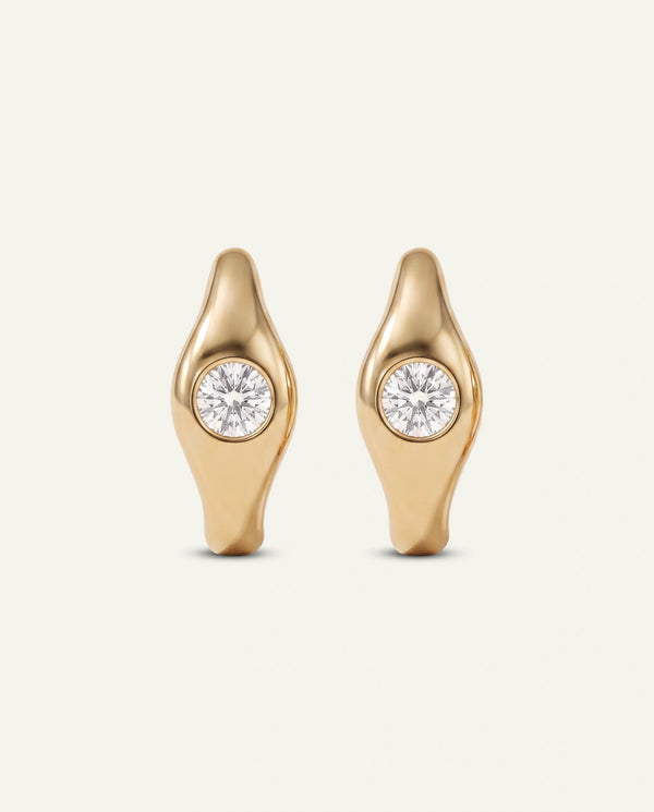 A pair of gold earrings with an organic, elongated teardrop shape and a round clear gemstone set in the center, shown against a light off-white background.