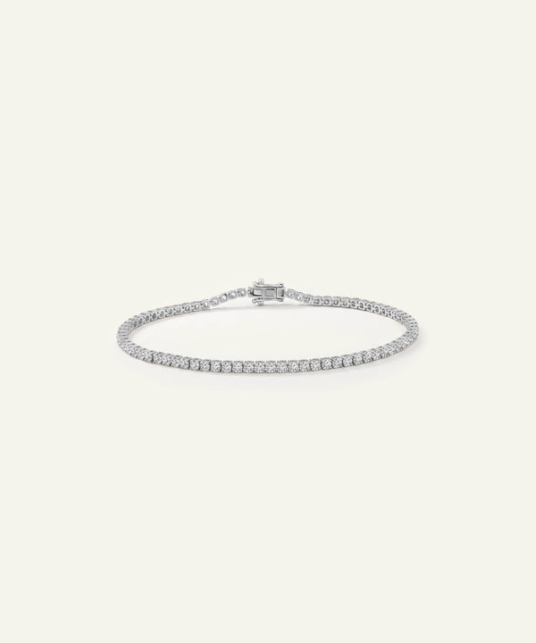 A silver tennis bracelet with a row of small clear stones, displayed on a light cream background.