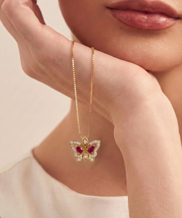 A close-up lifestyle shot of a model wearing a delicate gold chain necklace with a butterfly pendant. The butterfly is adorned with small clear crystals and vibrant red gemstones, resting against the model's neck.