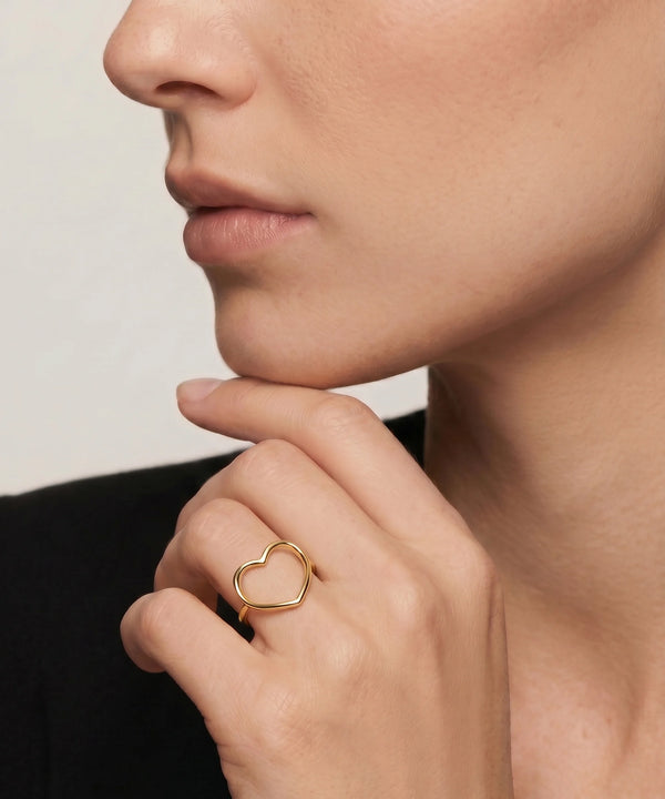 Close-up of a person wearing a gold heart-shaped ring on a neutral background