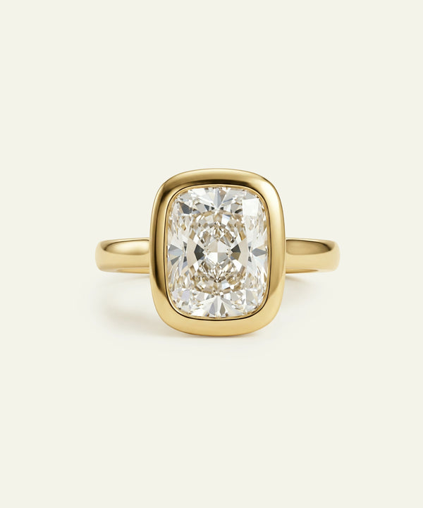 A gold ring featuring a large cushion-cut diamond in a bezel setting, shown against a light cream background.