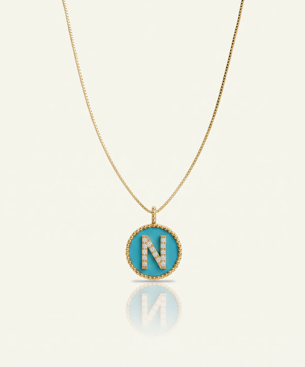 A gold chain necklace with an oval pendant featuring a turquoise blue background and a gold border, set with a large letter 'N' paved with small clear stones, shown on a light cream background with a soft reflection below.