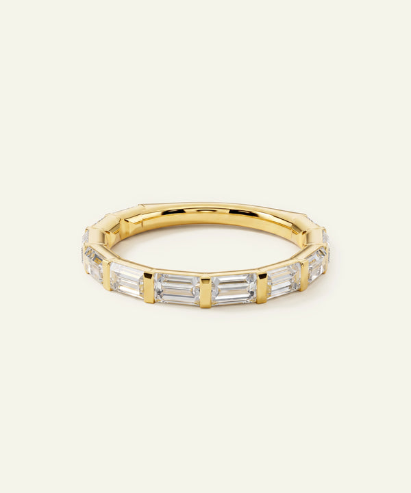 A gold band ring featuring a row of clear baguette-cut gemstones, photographed on a clean, solid off-white background.