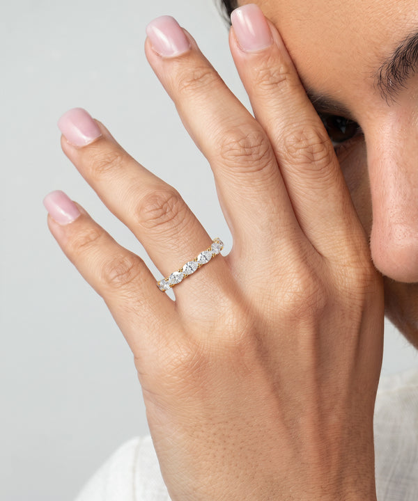 A close-up of a person's hand wearing a delicate silver band ring with a row of clear pavé-set gemstones, against a light gray background.