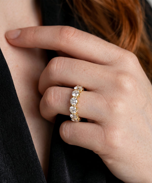 A close-up of a person's hand wearing a yellow gold eternity ring set with round brilliant-cut diamonds. The hand is resting against a black top, with reddish-brown hair visible in the background.