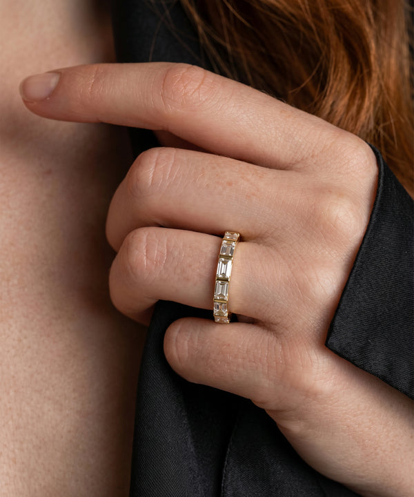 A close-up of a hand wearing a gold eternity ring with baguette-cut clear stones, set against a dark black shirt and warm skin tones.