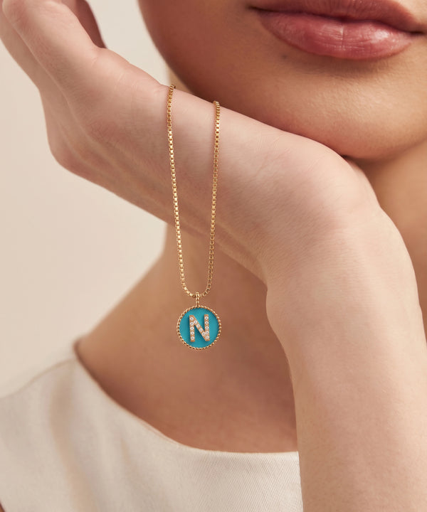 A close-up lifestyle shot of a model wearing a delicate gold chain necklace with a circular turquoise pendant featuring the letter 'N' in the center.