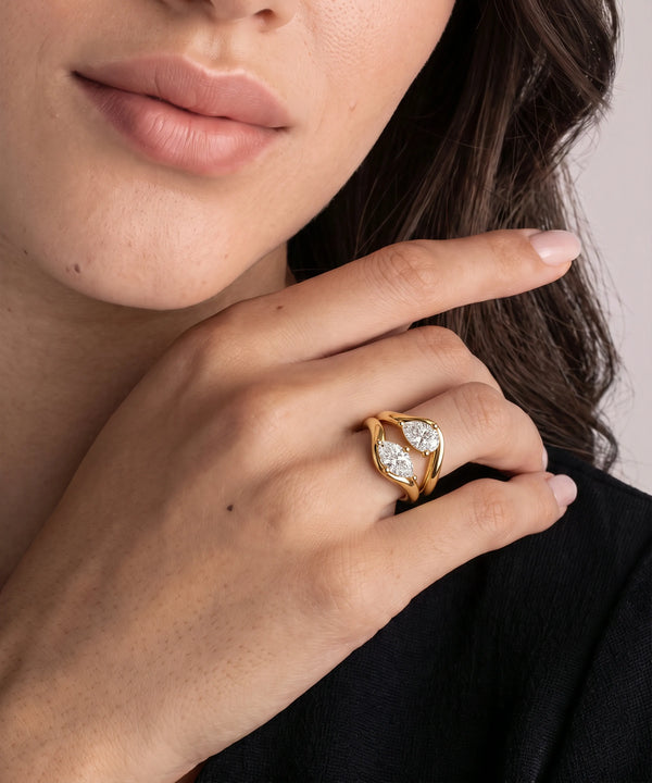 A close-up of a woman's hand wearing a gold ring with two marquise-cut clear gemstones in a leaf-like setting, against a soft neutral background.