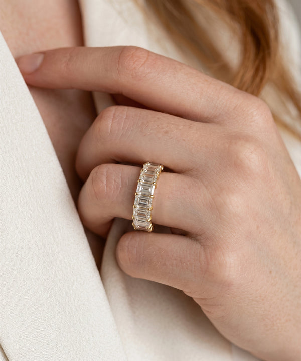 A close-up of a person's hand wearing a gold eternity ring with baguette-cut clear stones, set against a soft cream-colored coat.