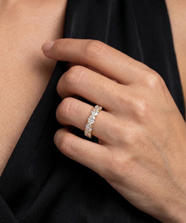 A close-up of a person's hand wearing a diamond eternity ring on a gold band, set against a black velvet garment.