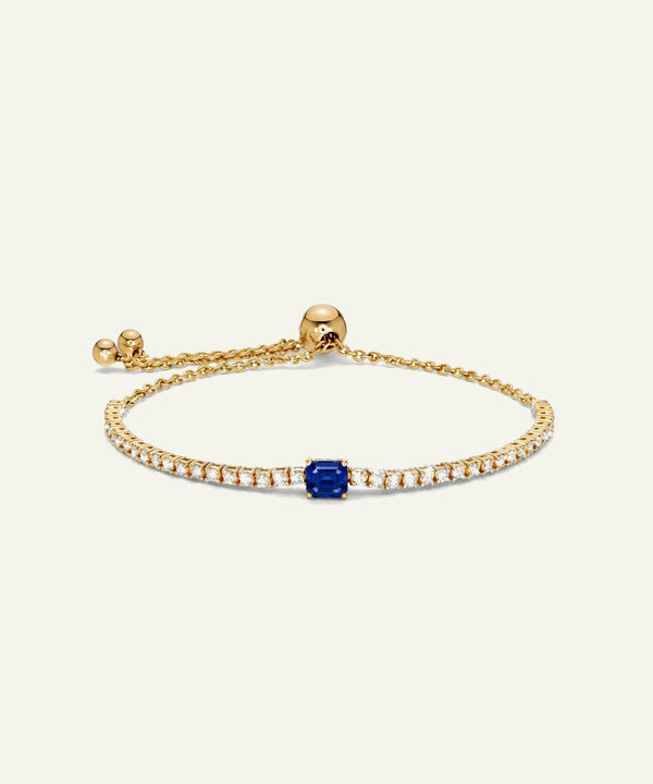 A delicate gold chain bracelet featuring a central square-cut blue sapphire surrounded by small clear stones, displayed on a clean off-white background.