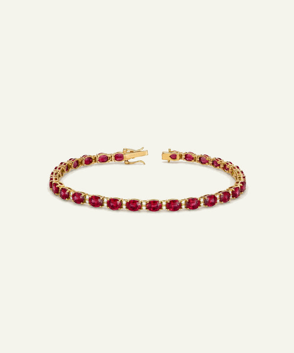 A gold tennis bracelet featuring a continuous row of oval-cut deep red gemstones, displayed on a clean, solid off-white background.