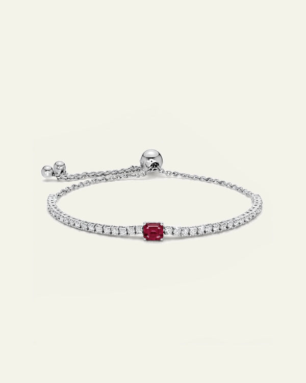 A silver chain bracelet featuring a single central red gemstone and a row of small clear stones along the top curve, shown on a plain light beige background.