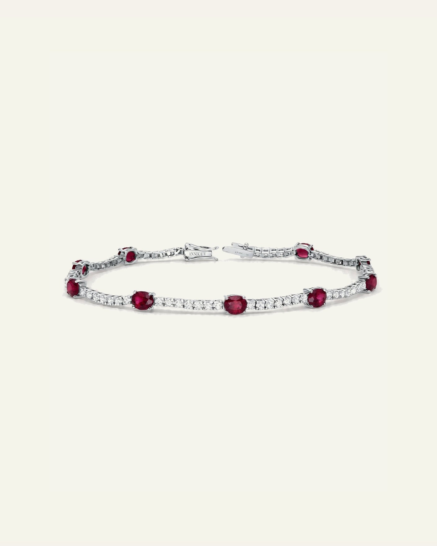 A silver tennis bracelet featuring oval red gemstones and rows of small clear stones, displayed on a light cream background.