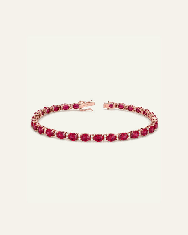 A rose gold tennis bracelet featuring a continuous line of oval-cut deep pinkish-red gemstones, displayed on a clean, solid off-white background.