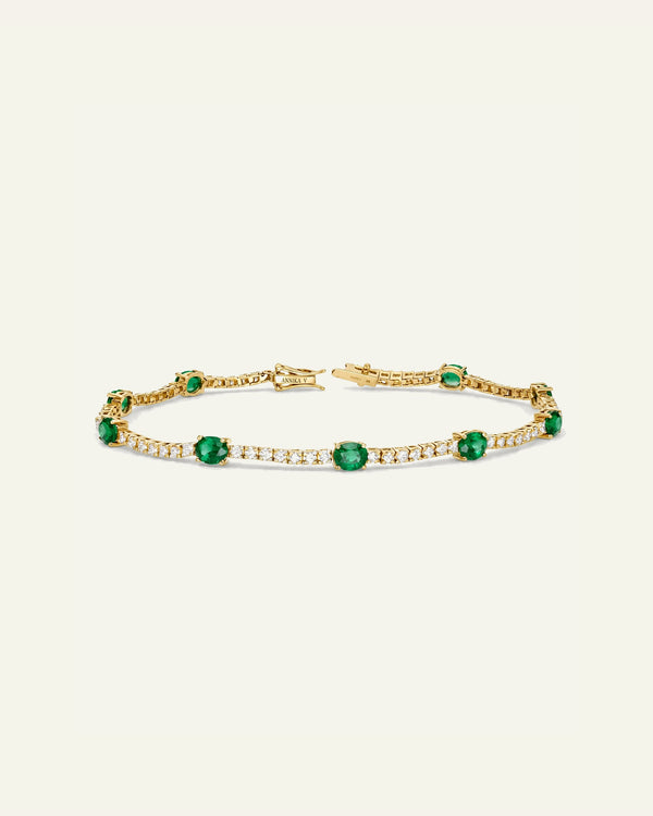 A gold tennis bracelet featuring round-cut green emeralds and rows of small clear stones, displayed on a light cream background.