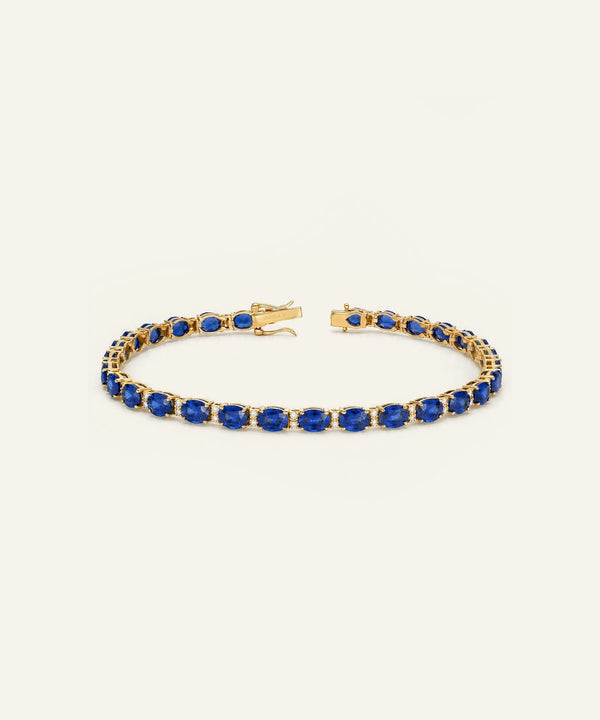 A gold tennis bracelet featuring a row of deep blue oval-cut gemstones, displayed on a clean, light off-white background.