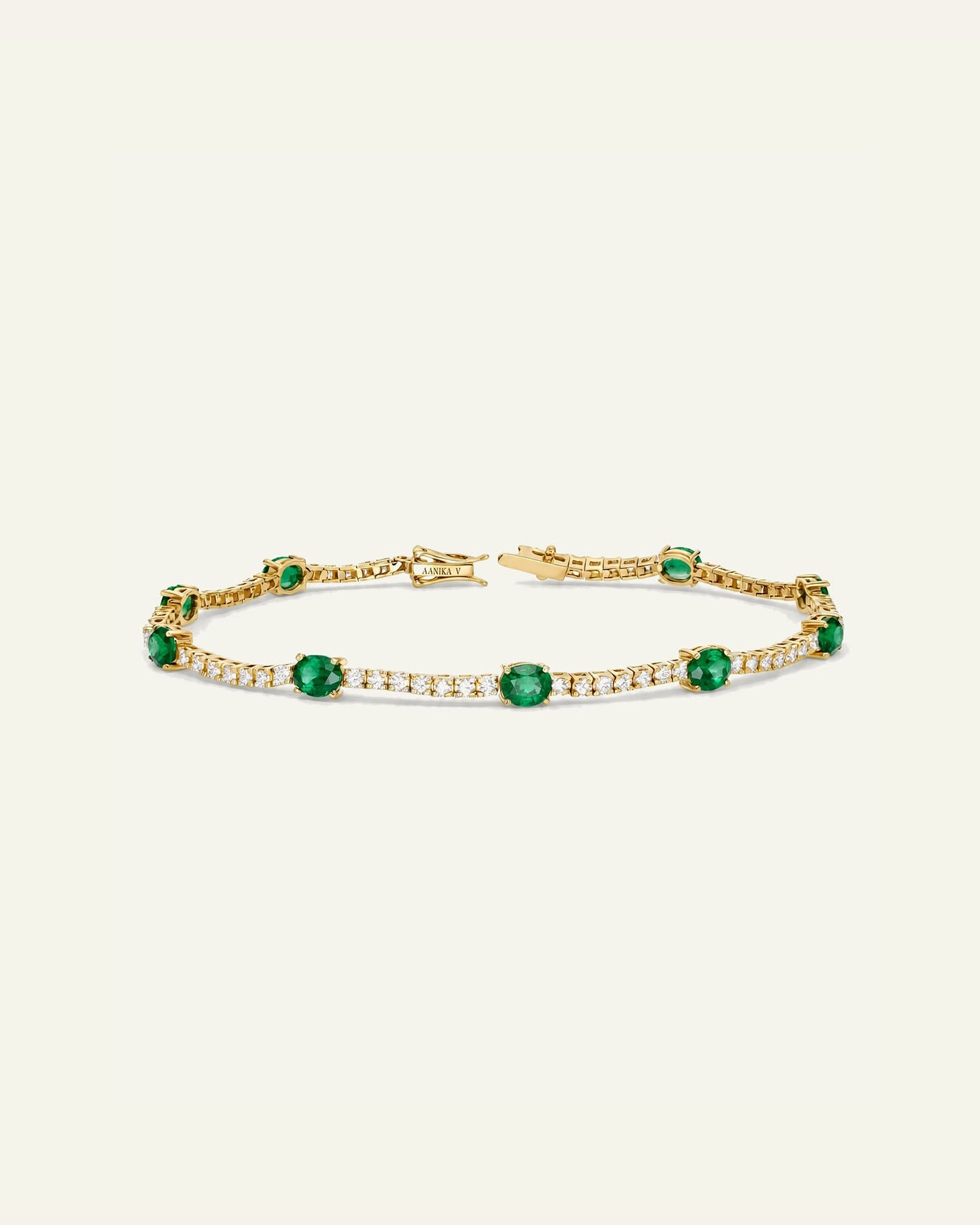 A gold tennis bracelet featuring round-cut green emeralds and rows of small clear stones, displayed on a light cream background.