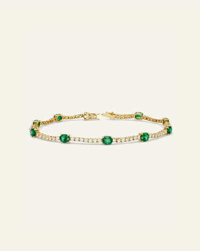 A gold tennis bracelet featuring round-cut green emeralds and rows of small clear stones, displayed on a light cream background.