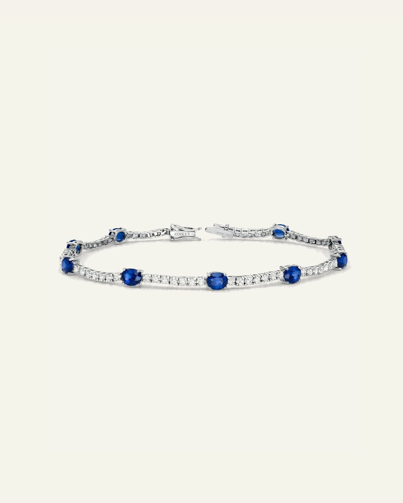 A silver tennis bracelet featuring round-cut blue sapphires and pavé-set clear stones, displayed on a solid cream background.