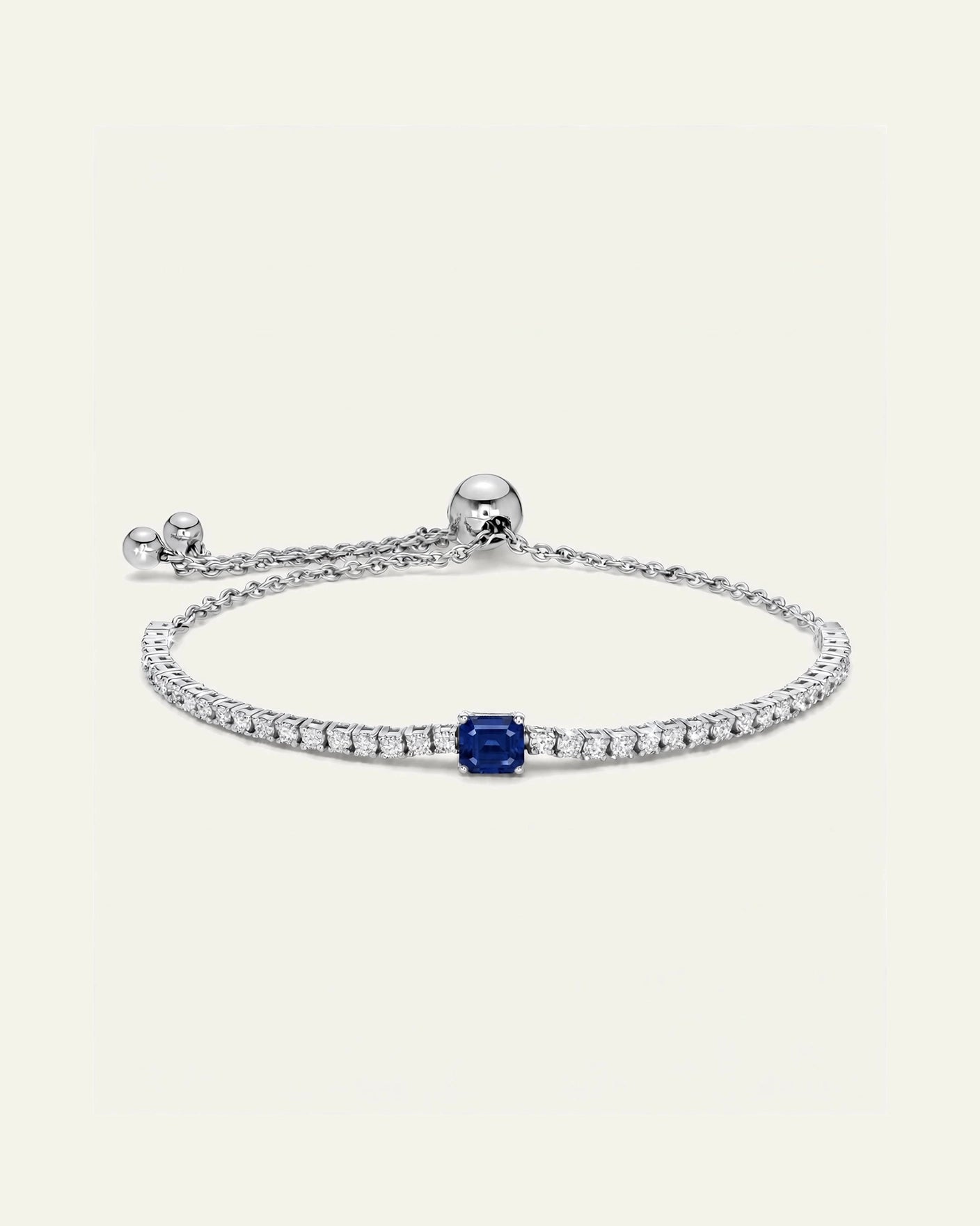 A delicate silver bracelet featuring a row of small clear stones, a central square-cut blue sapphire, and an adjustable sliding chain clasp, set against a solid off-white background.
