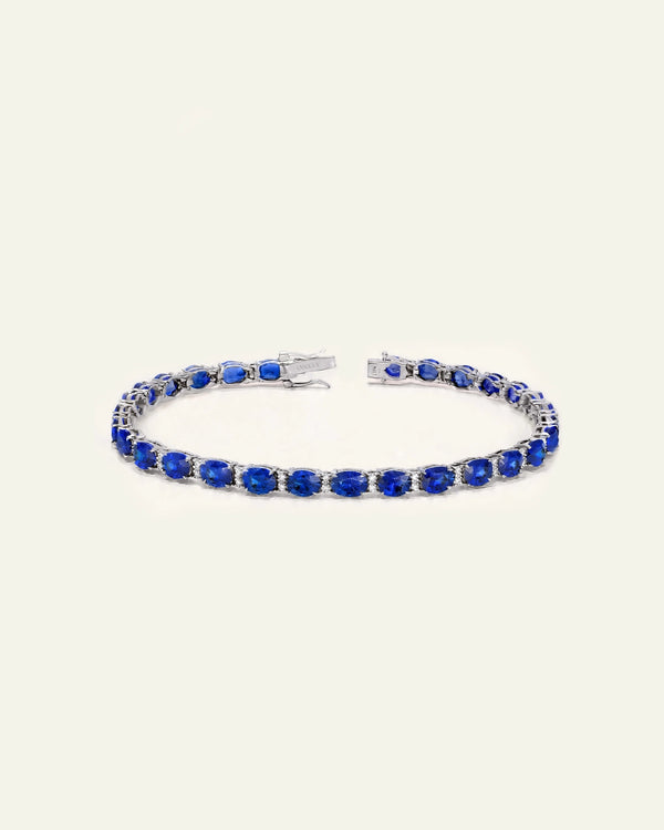 A silver-toned tennis bracelet featuring a row of oval-cut deep blue sapphires, displayed on a clean, solid off-white background.