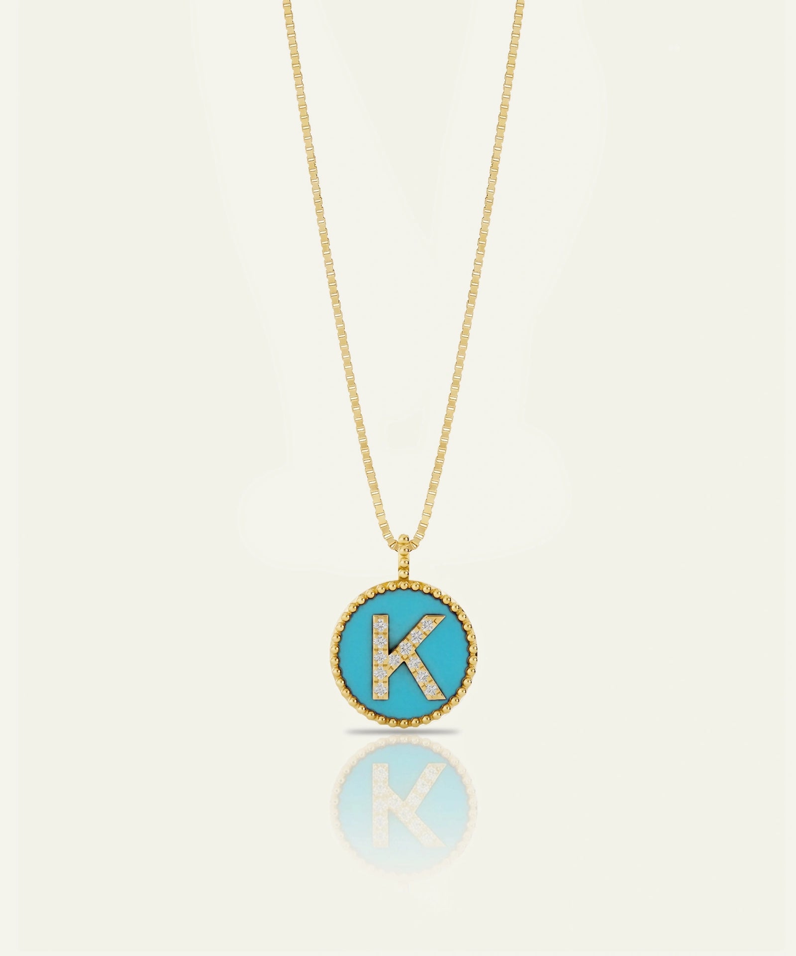A gold chain necklace with a round pendant featuring a turquoise blue background and a raised gold letter 'K' outlined with small clear stones, shown on a light cream background with a soft reflection below.